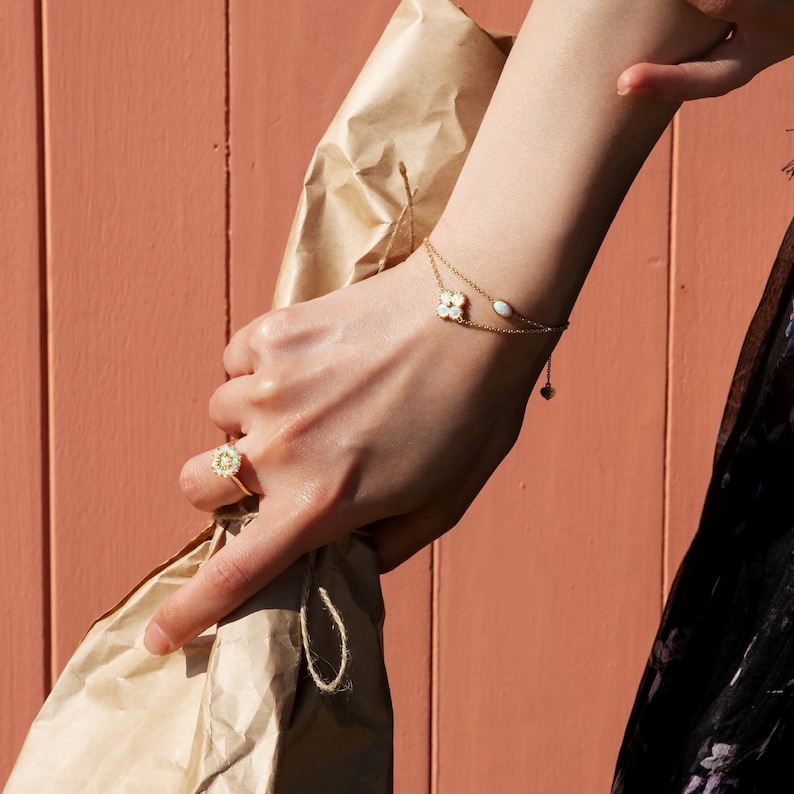 May include: A woman's hand wearing a gold ring and a delicate gold bracelet with a white flower charm and a small oval charm. The bracelet is on a brown paper bag.