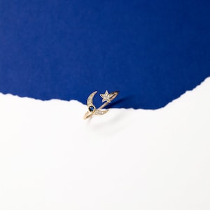 May include: A gold-colored ring featuring a crescent moon and a star design. The moon has a small blue gemstone and is adorned with small white gemstones. The star is also embellished with white gemstones. The ring is set against a blue and white background.