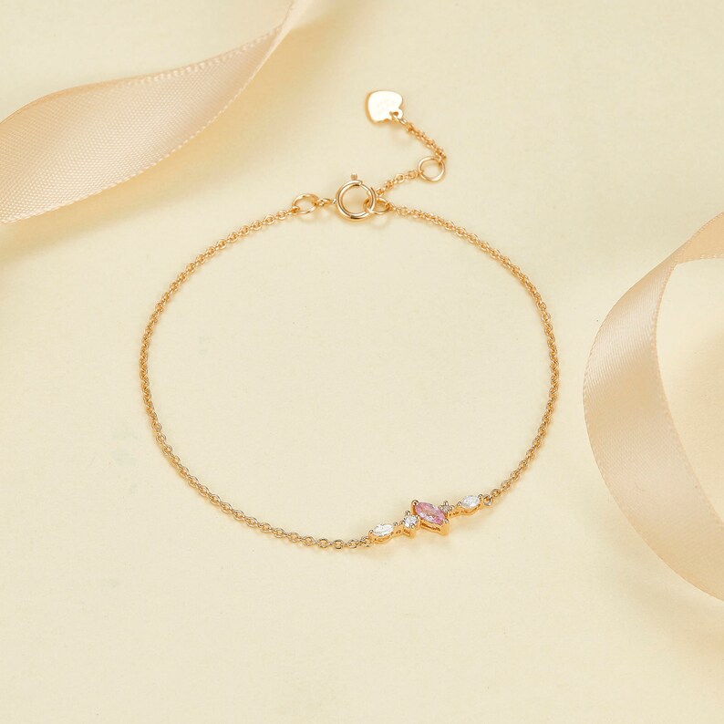 May include: A delicate gold chain bracelet with a pink and white gemstone cluster. The bracelet has a heart-shaped lobster clasp.