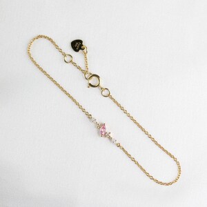 May include: A delicate gold chain bracelet with a pink oval gemstone and smaller clear gemstones. The bracelet has a heart-shaped clasp with the word "Cutie" engraved on it.
