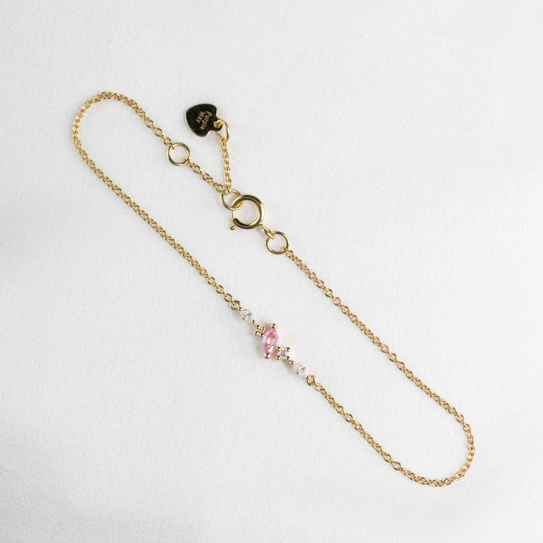May include: A delicate gold chain bracelet with a pink oval gemstone and smaller clear gemstones. The bracelet has a heart-shaped clasp with the word "Cutie" engraved on it.