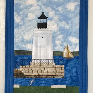 May include: A white lighthouse with a black roof stands on a stone pier. The lighthouse is surrounded by blue water and a sailboat is in the distance. The sky is blue with white clouds.