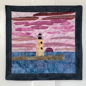 May include: A quilted wall hanging featuring a lighthouse on a small island with a pink and purple sunset in the background.