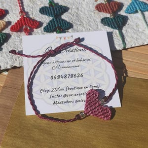 May include: A burgundy macrame bracelet with a heart charm. The bracelet is on a white card with the text "Créations Bijoux artisanaux et bohèmes Micromacramé 0684878626 Etsy: ZDCox (boutique en ligne) Insta: @cox-creatic Mastodon: @ciss".