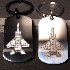 May include: Two silver keychains with black key rings. Each keychain features a dog tag with an engraved image of a fighter jet.