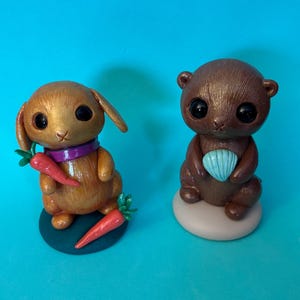 May include: Two handcrafted figurines: a golden-brown bunny with a purple scarf and carrots, and a brown otter holding a blue seashell. Both have large, dark eyes. The bunny sits on a green base, the otter on a beige one, against a blue backdrop.