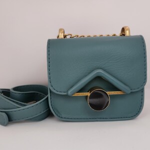May include: A small teal blue crossbody bag with a gold chain strap and a circular gold accent with a black center.