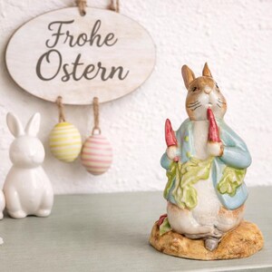 Könnte beinhalten: Osterdekoration mit einer Keramik-Hasenfigur, die Karotten hält, einem Schild mit der Aufschrift "Frohe Ostern" und dekorativen Eiern. Eine Vase mit gelben Tulpen und weißen Keramikhasen vervollständigen die Frühlingsdekoration.