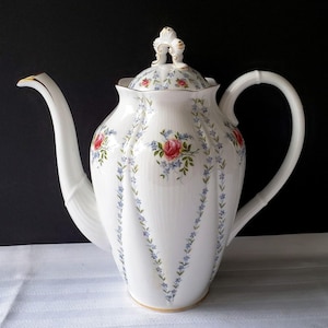 Pode incluir: Bule de porcelana branco com um design floral de rosas vermelhas e flores azuis. O bule tem um bico curvo, uma alça arredondada e uma tampa decorativa com detalhes dourados. Em estilo vintage.