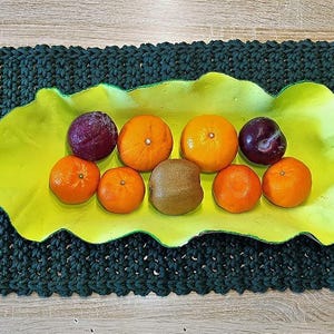 May include: A dark green knitted placemat with a bright yellow, leaf-shaped bowl filled with fresh fruit. The bowl holds oranges, plums, and a kiwi. The placemat is rectangular and has a textured surface.