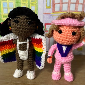 May include: Two crocheted dolls, one with brown skin and a rainbow cape, the other with peach skin and a pink suit.