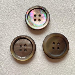 May include: Three round buttons, each with four holes. The top button has an iridescent surface with green, purple, and gold hues. The other two buttons are a solid brown colour.
