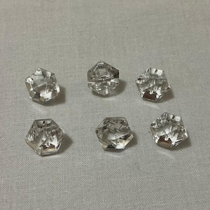May include: Six clear, faceted crystal beads with a hexagonal shape are arranged on a light grey fabric. The beads have multiple reflective surfaces, catching the light and creating a sparkling effect. These beads are suitable for jewellery making or craft projects.