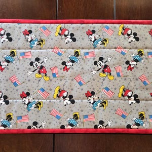 May include: A rectangular quilted table runner featuring a light gray fabric background with a repeating pattern of Mickey and Minnie Mouse characters, small dark gray stars, and miniature American flags, finished with a solid red fabric border.