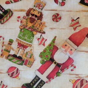 K&ouml;nnte beinhalten: Ein wei&szlig;er Stoff mit Weihnachtsmotiv mit Lebkuchenmann, Nussknacker, Weihnachtsmann, Zuckerstangen und Ornamenten. Der Stoff hat einen Holzmaserungshintergrund.