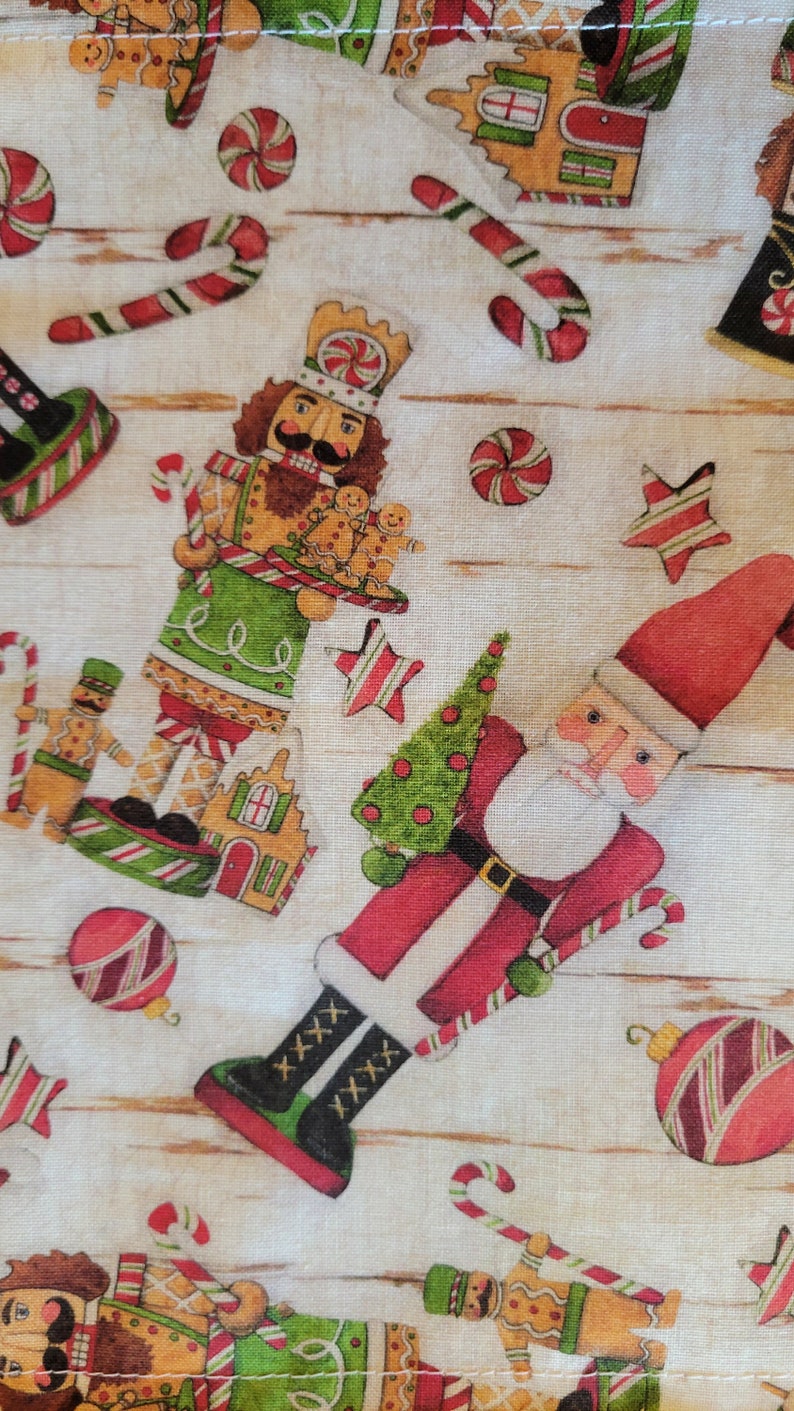 K&ouml;nnte beinhalten: Ein wei&szlig;er Stoff mit Weihnachtsmotiv mit Lebkuchenmann, Nussknacker, Weihnachtsmann, Zuckerstangen und Ornamenten. Der Stoff hat einen Holzmaserungshintergrund.