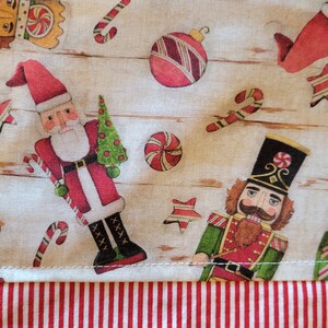 K&ouml;nnte beinhalten: Ein wei&szlig;er Stoff mit einem rot-wei&szlig; gestreiften Rand. Der Stoff zeigt ein sich wiederholendes Muster aus Nussknackern, Zuckerstangen, Kugeln und Sternen. Das Muster ist in Rot-, Gr&uuml;n-, Braun- und Wei&szlig;t&ouml;nen gehalten.