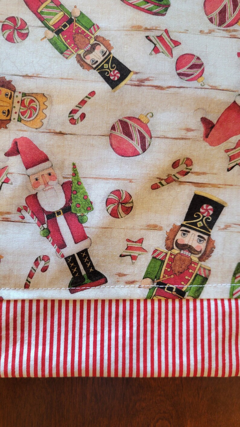 K&ouml;nnte beinhalten: Ein wei&szlig;er Stoff mit einem rot-wei&szlig; gestreiften Rand. Der Stoff zeigt ein sich wiederholendes Muster aus Nussknackern, Zuckerstangen, Kugeln und Sternen. Das Muster ist in Rot-, Gr&uuml;n-, Braun- und Wei&szlig;t&ouml;nen gehalten.