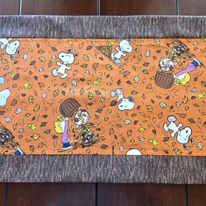 May include: A rectangular table runner featuring a central orange fabric panel printed with a repeating pattern of Snoopy, Woodstock, and Sally Brown playing in autumn leaves, bordered by a dark brown fabric with a textured wood grain print.