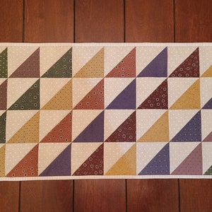 May include: A rectangular quilted table runner with a geometric pattern of alternating triangles. The triangles are in shades of green, brown, purple, and yellow, with a cream-colored background and a white border.