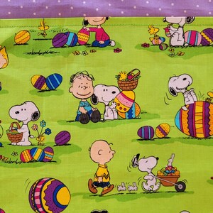 May include: A green fabric with a pattern of Peanuts characters celebrating Easter. The characters are Snoopy, Charlie Brown, and Woodstock. They are holding Easter eggs and baskets. The fabric has a purple border with white polka dots.