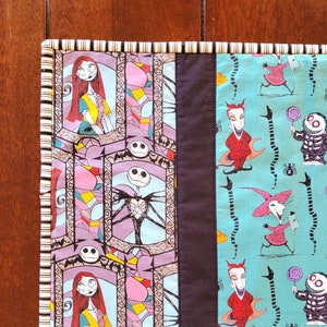 May include: A colorful patchwork quilt runner featuring characters from the Nightmare Before Christmas. The runner is made of different fabrics with various patterns, including a purple and pink geometric pattern, a blue and white striped pattern, and a turquoise pattern with black snakes and characters from the movie. The runner is trimmed with a white and black striped border.