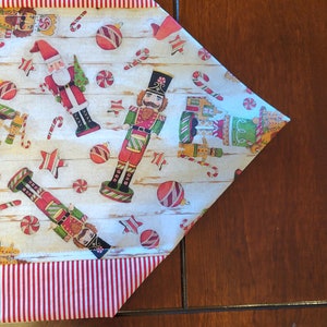 K&ouml;nnte beinhalten: Ein wei&szlig;er Tischl&auml;ufer mit rotem und wei&szlig;em Streifenrand. Der L&auml;ufer zeigt ein Weihnachtsmotiv mit Nussknacker-Soldaten, Zuckerstangen und Ornamenten.