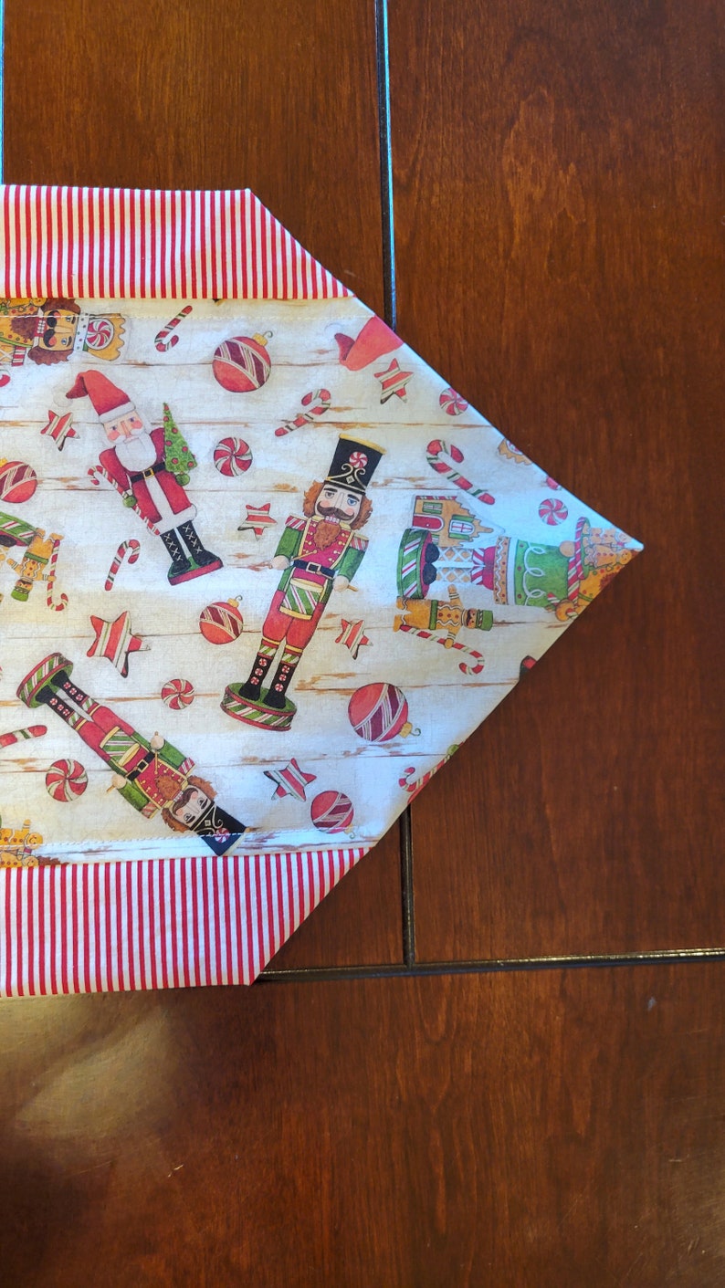 K&ouml;nnte beinhalten: Ein wei&szlig;er Tischl&auml;ufer mit rotem und wei&szlig;em Streifenrand. Der L&auml;ufer zeigt ein Weihnachtsmotiv mit Nussknacker-Soldaten, Zuckerstangen und Ornamenten.