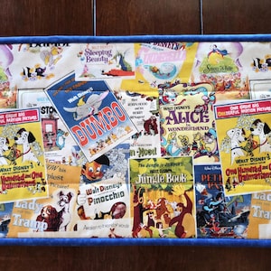 May include: A rectangular fabric runner featuring a collage of vintage Walt Disney movie posters including Dumbo, Alice in Wonderland, The Jungle Book, Pinocchio, Lady and the Tramp, and 101 Dalmatians, finished with a solid blue fabric border.