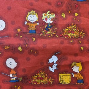 May include: A red fabric featuring a repeating pattern of Peanuts characters playing in autumn leaves. Charlie Brown, Snoopy, and other characters are depicted in a fall scene with piles of leaves and brooms. The fabric has a light tan border.