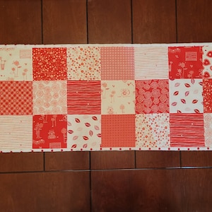 May include: A red and white patchwork table runner with a variety of patterns, including hearts, flowers, and geometric designs. The runner has a white border with red polka dots.