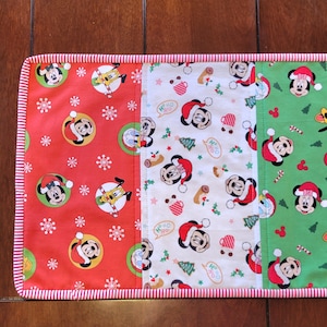May include: A red, white, and green Christmas table runner with a Mickey Mouse theme. The runner features a variety of Mickey Mouse designs, including Mickey Mouse wearing a Santa hat, Mickey Mouse with a candy cane, and Mickey Mouse with a Christmas tree. The runner is trimmed with a red and white striped border.