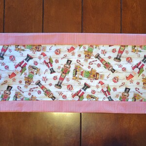 K&ouml;nnte beinhalten: Ein wei&szlig;er Tischl&auml;ufer mit einem rot-wei&szlig; gestreiften Rand. Der L&auml;ufer zeigt ein sich wiederholendes Muster aus Nussknackerfiguren, Zuckerstangen und Kugeln.