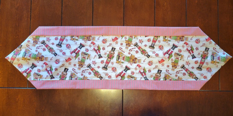 K&ouml;nnte beinhalten: Ein wei&szlig;er Tischl&auml;ufer mit einem rot-wei&szlig; gestreiften Rand. Der L&auml;ufer zeigt ein sich wiederholendes Muster aus Nussknackerfiguren, Zuckerstangen und Kugeln.