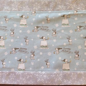 May include: A light blue fabric table runner featuring a repeating pattern of Snoopy wearing a winter hat, Snoopy with antlers, and the text Happy Holidays, interspersed with white snowflakes. The runner has wide borders with a white snowflake print.