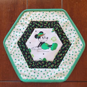 May include: A green, black, and gold hexagonal quilted table runner with a white center featuring a cartoon image of Mickey Mouse wearing a green hat and holding a shamrock. The runner is decorated with green shamrock patterns.