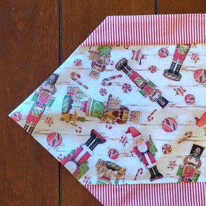 K&ouml;nnte beinhalten: Ein wei&szlig;er Tischl&auml;ufer mit einem rot-wei&szlig; gestreiften Rand. Der L&auml;ufer zeigt ein sich wiederholendes Muster aus Nussknackern, Zuckerstangen und Kugeln.