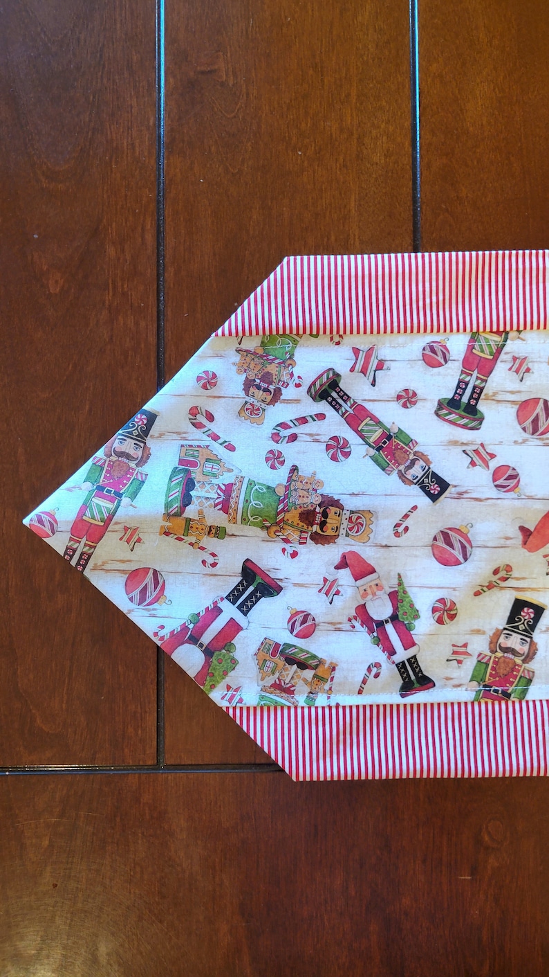 K&ouml;nnte beinhalten: Ein wei&szlig;er Tischl&auml;ufer mit einem rot-wei&szlig; gestreiften Rand. Der L&auml;ufer zeigt ein sich wiederholendes Muster aus Nussknackern, Zuckerstangen und Kugeln.