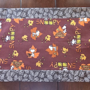 May include: A rectangular fabric table runner featuring a brown center panel with a repeating pattern of Snoopy, autumn leaves, and the text SNOOPY. The runner has dark brown borders on the top and bottom edges decorated with a white leaf print.