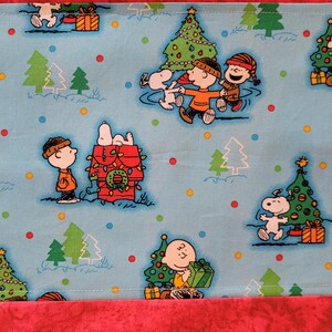 May include: A blue fabric with a repeating pattern of Peanuts characters celebrating Christmas. The characters include Snoopy, Charlie Brown, Lucy, and Linus. The fabric features a blue background with red, green, yellow, and white accents.