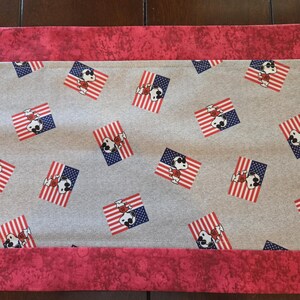 May include: A gray fabric table runner with a repeating pattern of Snoopy dressed in patriotic attire on American flags. The runner has a red border with a mottled design.