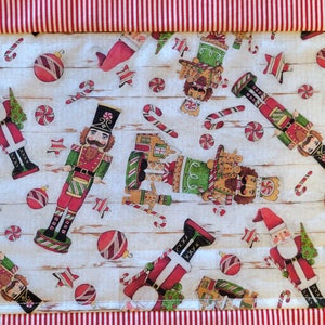 May include: A white fabric table runner with a red and white striped border. The runner features a repeating pattern of nutcrackers, candy canes, ornaments, and gingerbread men.