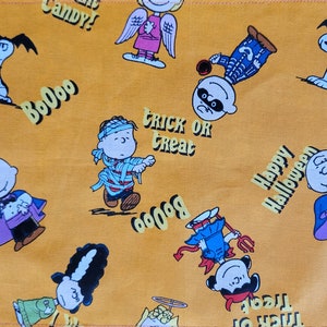 May include: A yellow fabric with a pattern of Peanuts characters dressed in Halloween costumes. The characters include Charlie Brown, Lucy, Linus, and Snoopy. The fabric also has the text "Boo!" and "Trick or Treat".