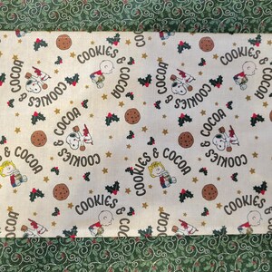 May include: A white fabric table runner with a green border. The runner features a repeating pattern of cartoon characters holding cookies and mugs, with the text "Cookies & Cocoa" repeated throughout. The background is a cream color with gold stars.