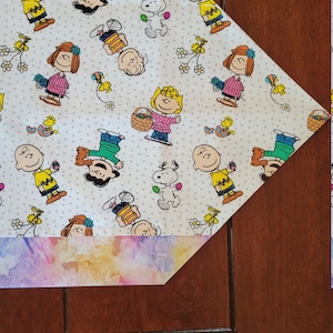 May include: A white table runner with a colorful watercolor border and a repeating pattern of Peanuts characters, including Snoopy, Charlie Brown, and Lucy. The characters are depicted in various poses, including holding Easter eggs and baskets.