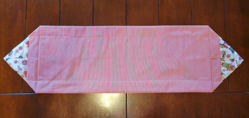 K&ouml;nnte beinhalten: Ein rot-wei&szlig; gestreifter Tischl&auml;ufer mit wei&szlig;em Stoffrand und einem festlichen Weihnachtsdruck an den Enden.