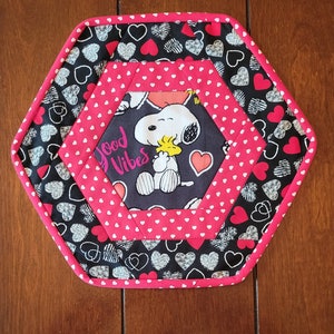 May include: Hexagon-shaped hot pad with a black background and white hearts. The center features a pink and white fabric with a white polka dot pattern. A cartoon image of Snoopy and Woodstock is in the center with the text "Good Vibes".