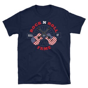 Op de afbeelding: Een marineblauw t-shirt met een witte afbeelding van twee gitaren die over elkaar heen gekruist zijn. De gitaren zijn versierd met rood-wit-blauwe strepen. De tekst "Rock N Roll Hall Of Fame" is in wit rond de gitaren gedrukt.