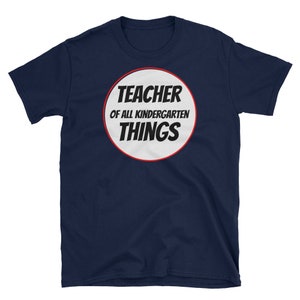 May include: Navy blue t-shirt with a white circle and red outline. The circle has the text "Teacher of all kindergarten things" in black.