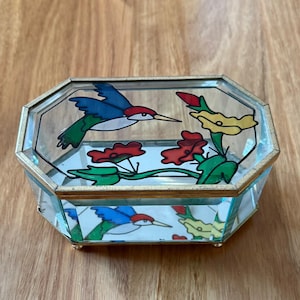 May include: An octagonal glass jewellery box with a gold-coloured metal frame. The box features a painted design of hummingbirds, red flowers, and yellow flowers. The box is sitting on a wooden surface.